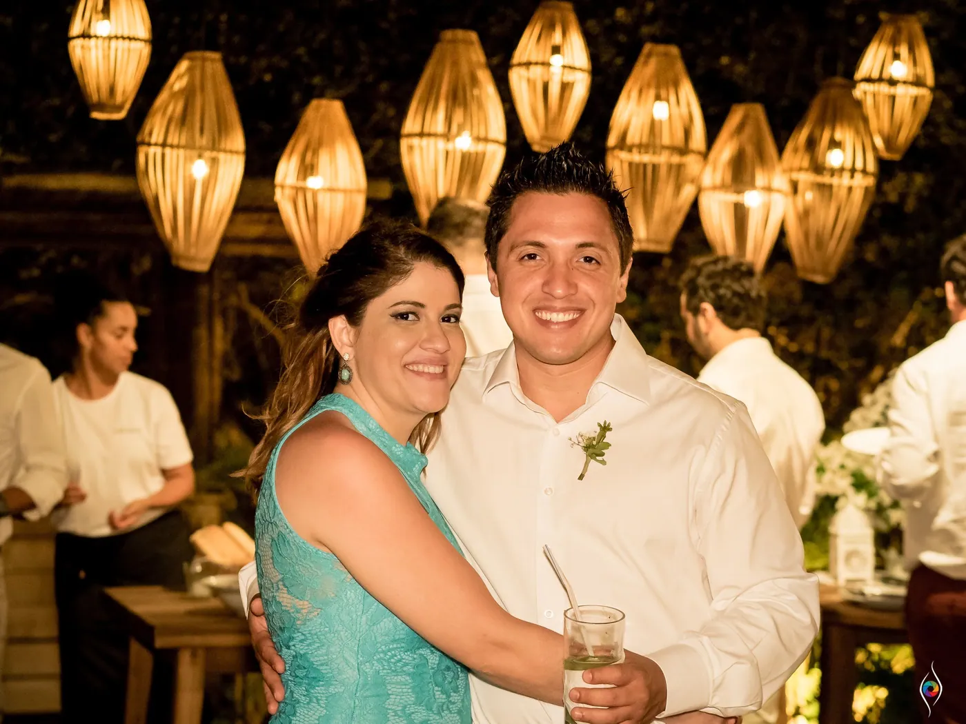 A couple hug close beneath hanging woven lamps, his boutonniere still pinned on