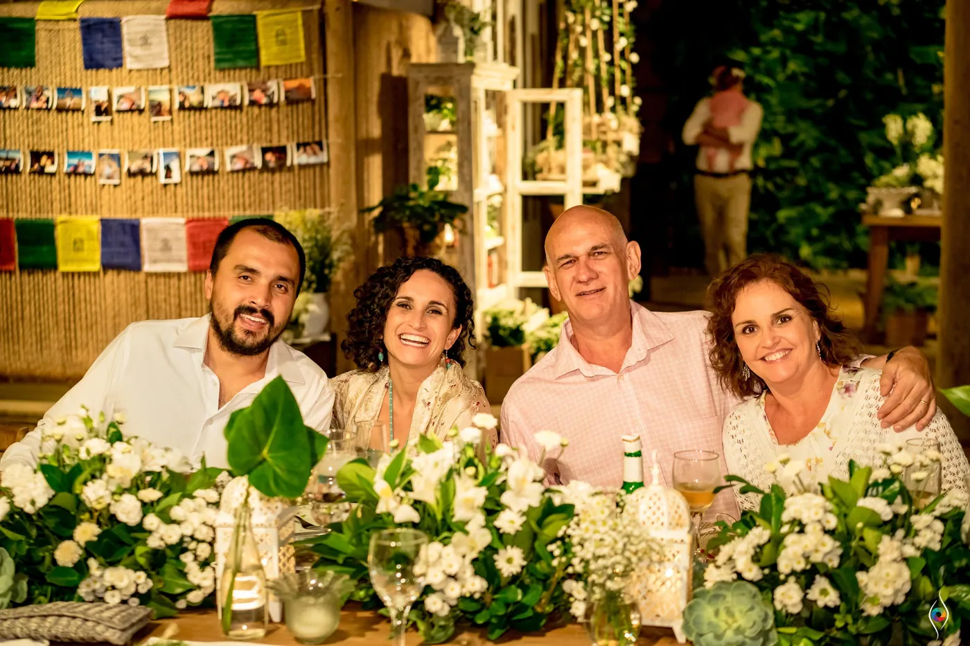 Four guests laugh at a table dressed in white flowers, a photo wall and flags behind them