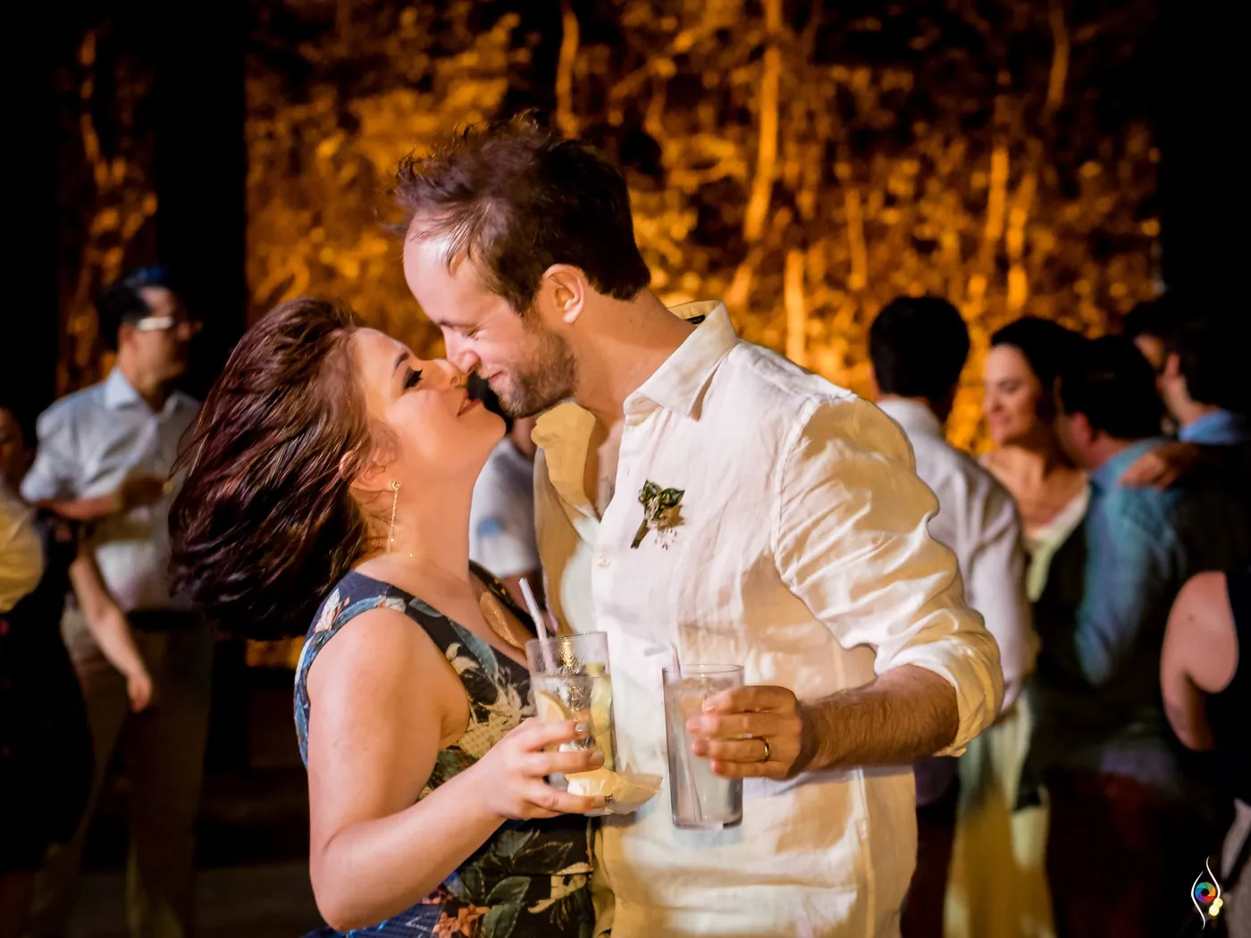 A woman in a navy floral dress kisses a man in white on the cheek, cocktails in hand