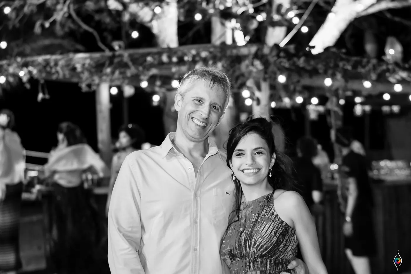 A man in a light shirt and a woman in a sleeveless dress smile under string lights