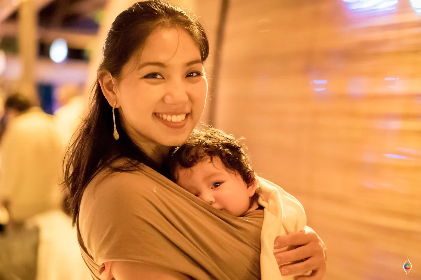 A woman in a taupe wrap smiles with a baby tucked against her chest in warm party light