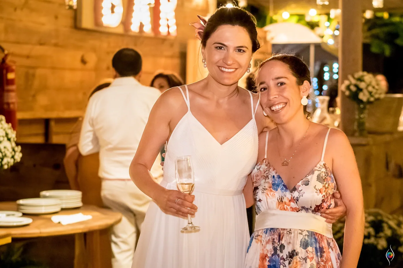 Evy raises a glass of sparkling wine beside a smiling woman in a floral dress at the lit bar
