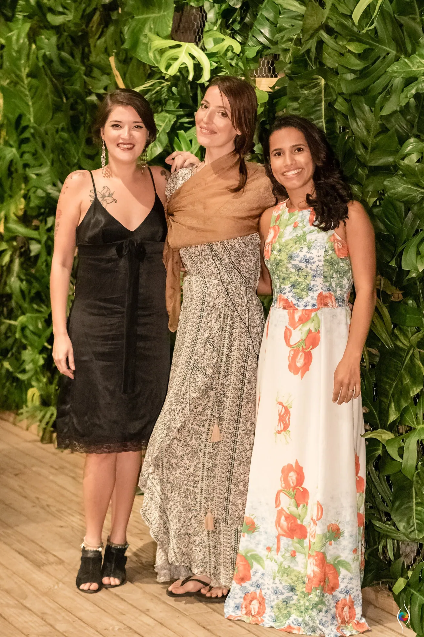 Three women in black, a shawl and a long floral dress pose in front of the leaf wall