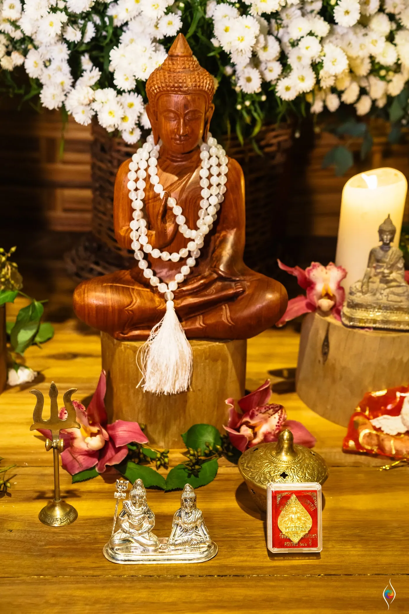 A carved Buddha statue with a shell necklace sits among orchids, a candle and small brass figures