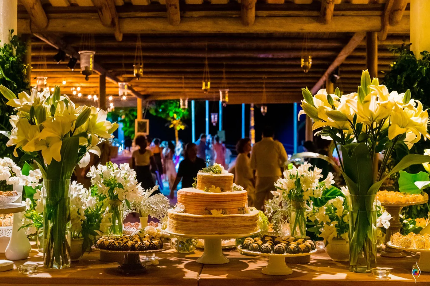 A tiered cake and tall yellow lilies glow on a table lit from above