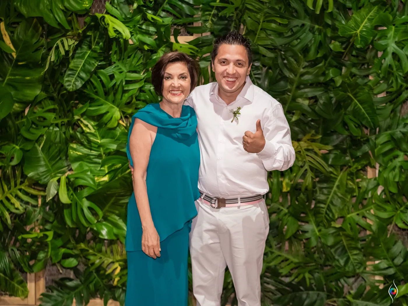 A woman in teal drapes stands beside a man in white who gives a thumbs up against green leaves