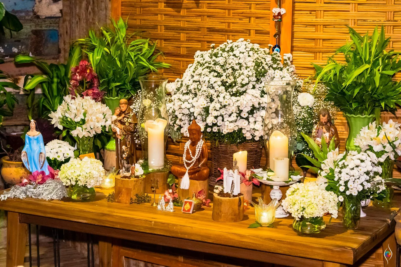 A candlelit table of white daisies and small religious figurines glows against a bamboo wall