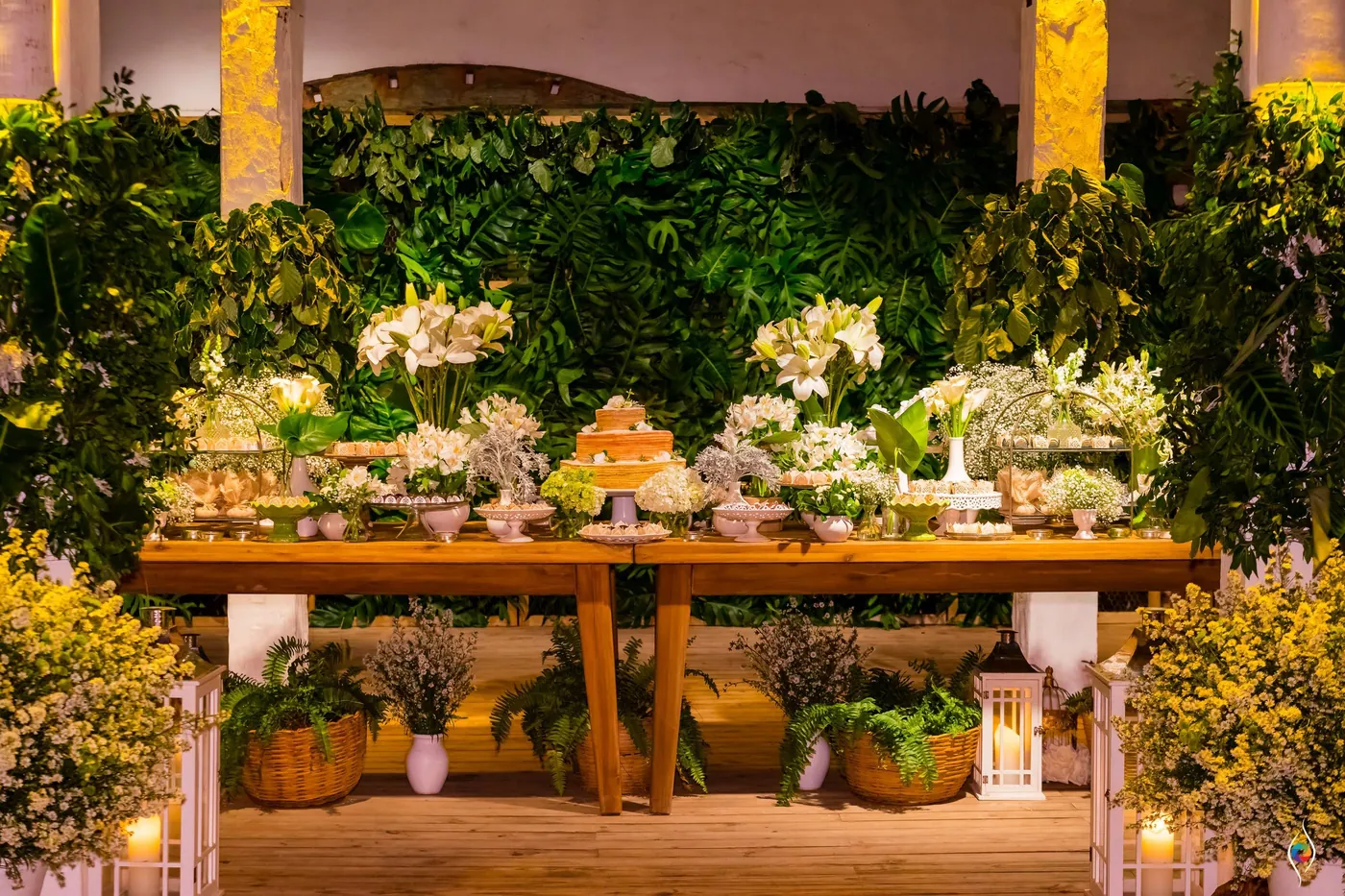 A long wooden table of flowers and candles stands before a wall of green