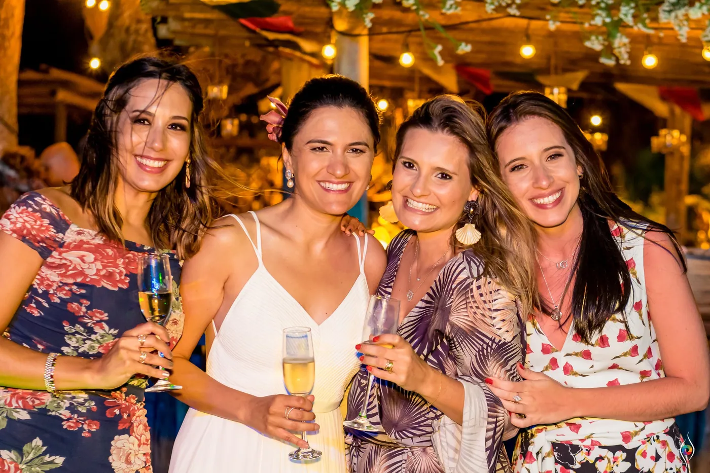 Evy in white holds a champagne flute, cheek to cheek with three friends in floral dresses