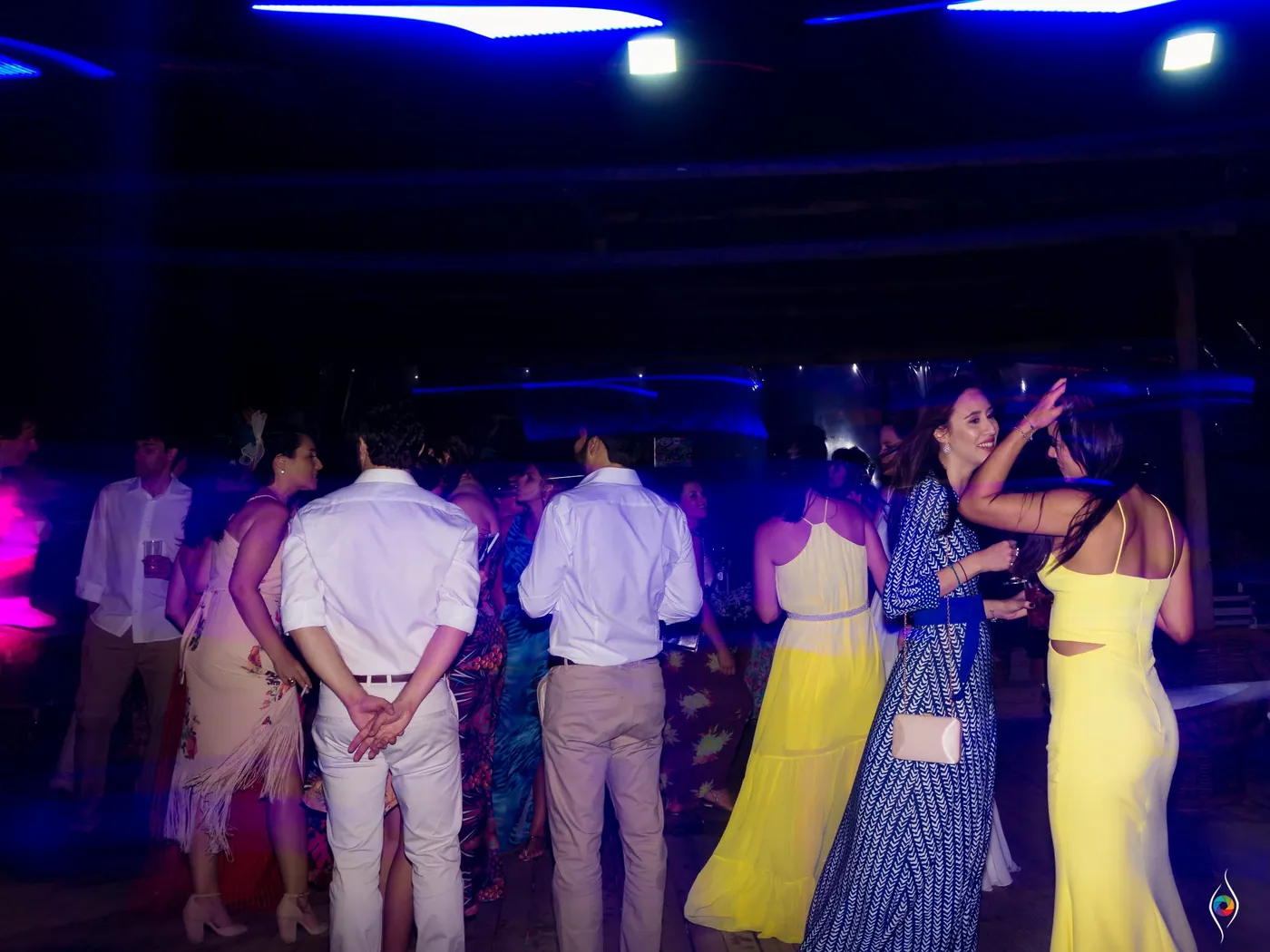 Guests in bright dresses dance under blue and violet stage light, backs to the camera