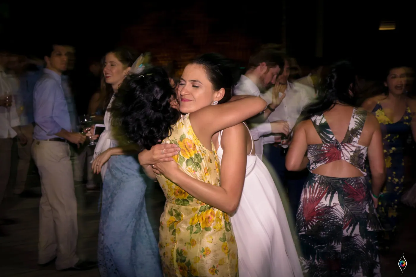 Evy hugs a friend in a yellow floral dress on the dance floor while guests dance behind