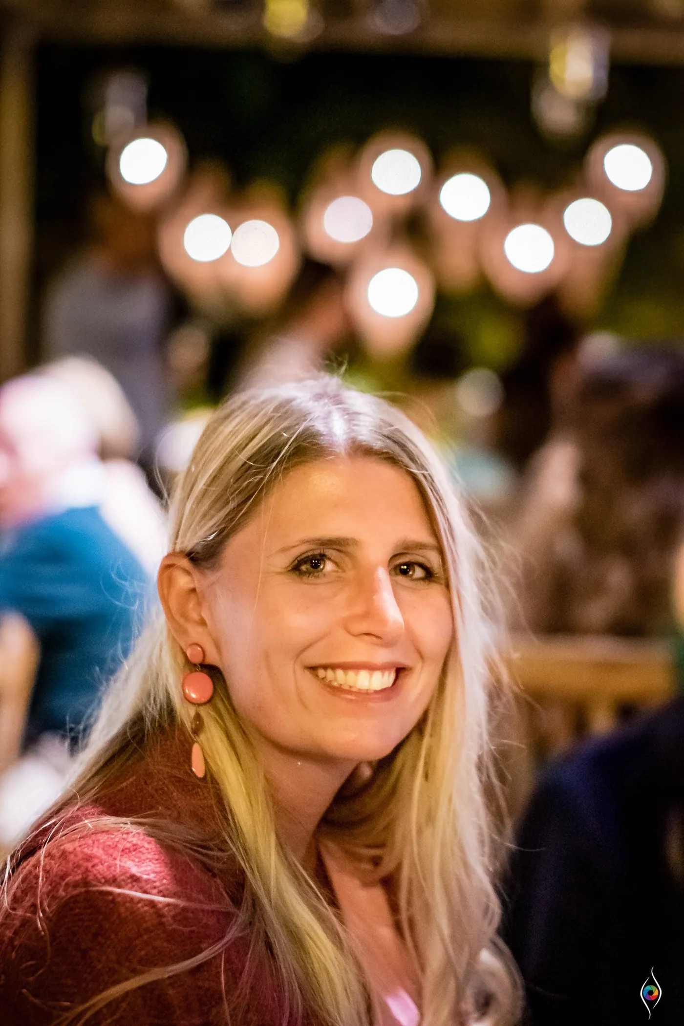 A blonde woman in a berry-coloured top smiles over her shoulder, string lights blurred behind her