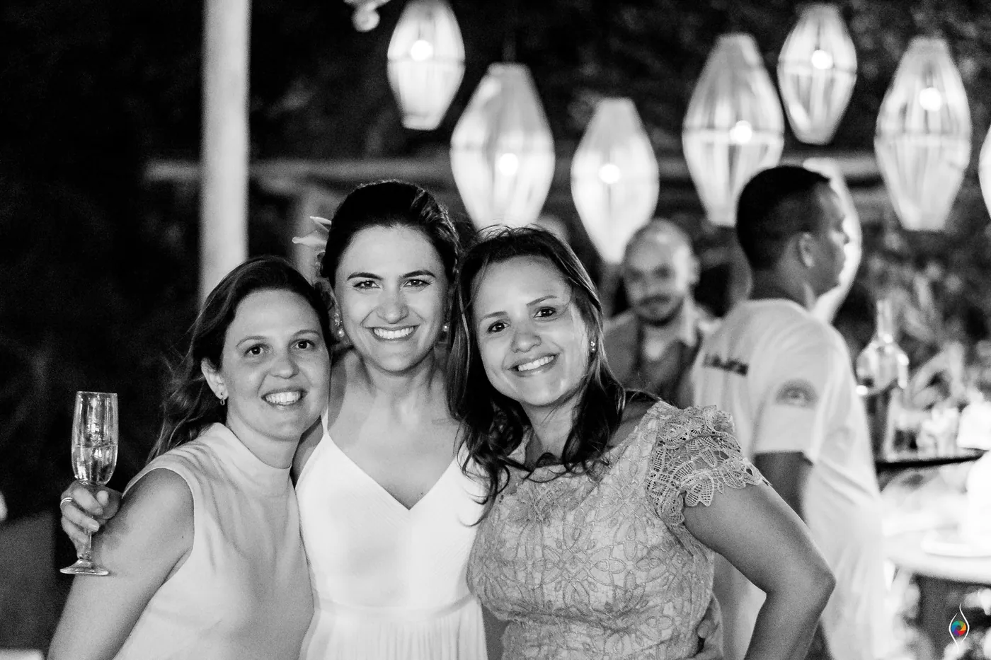 Evy in white poses with two friends, one holding a champagne flute, lanterns hanging behind