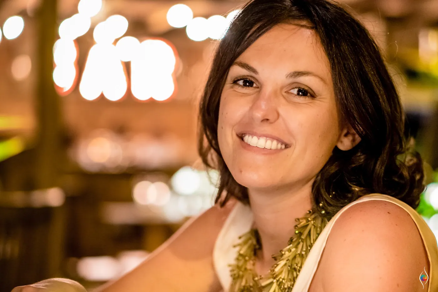 A woman with a gold necklace smiles straight at the camera against warm blurred lights