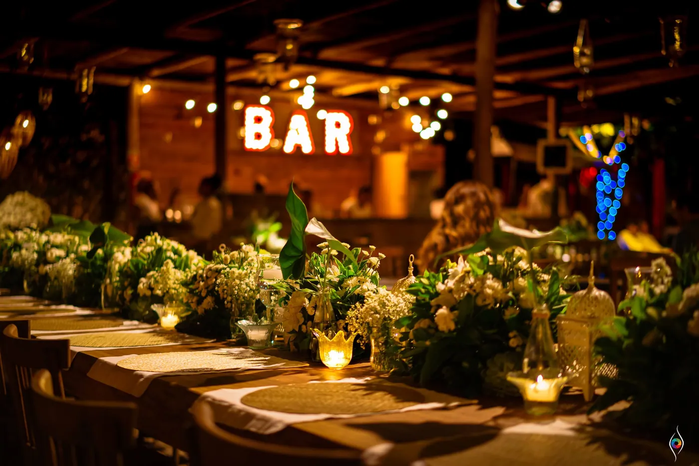 The long dinner table stretches toward a glowing BAR sign under the party canopy