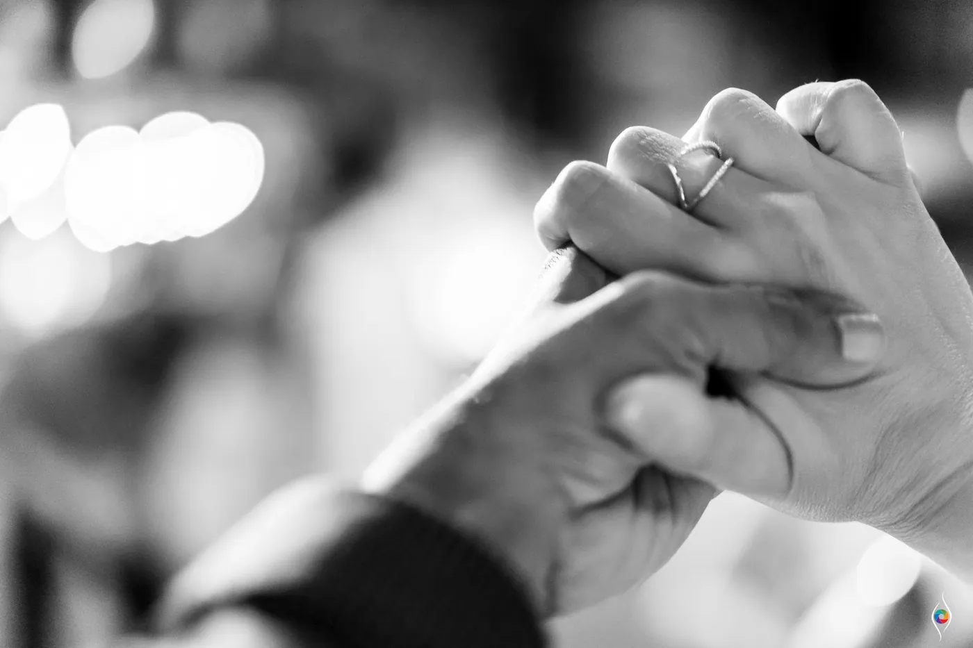 Clasped hands in black and white with a ring on one finger, soft lights blurred behind