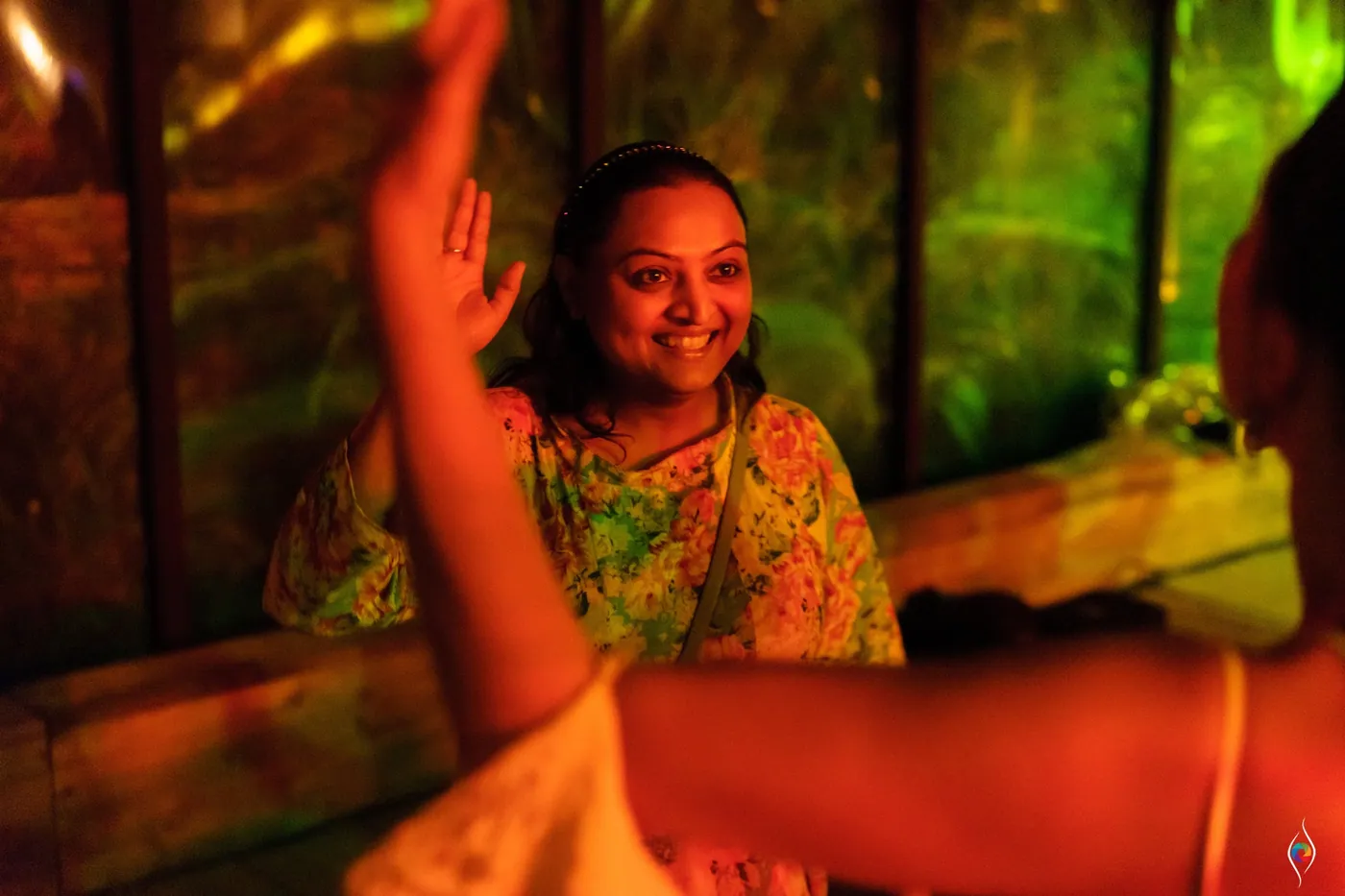 A smiling woman in a floral top sits as a dancer reaches an arm up in orange and green light